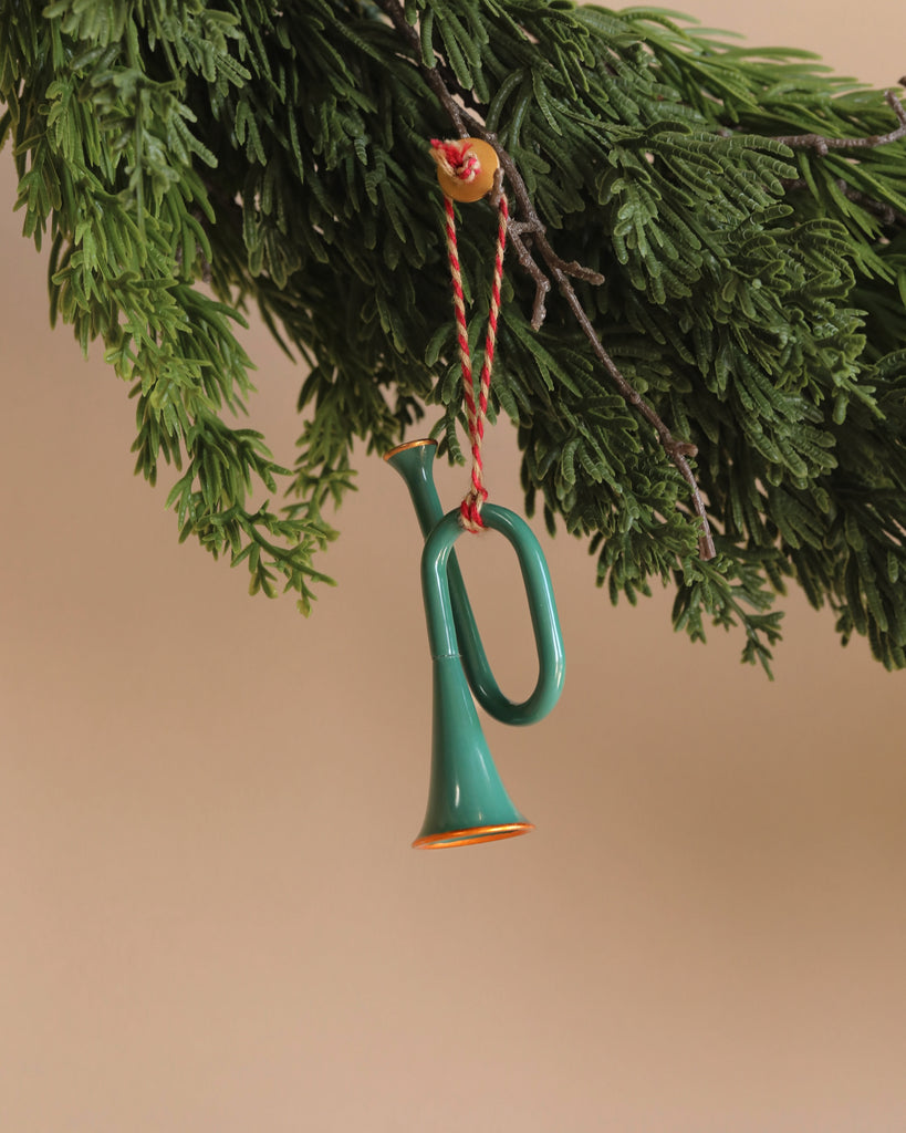 A Maileg Metal ornament, Trumpet - Dark Green with handpainted details and a red and white string hangs from a pine branch against a beige background.