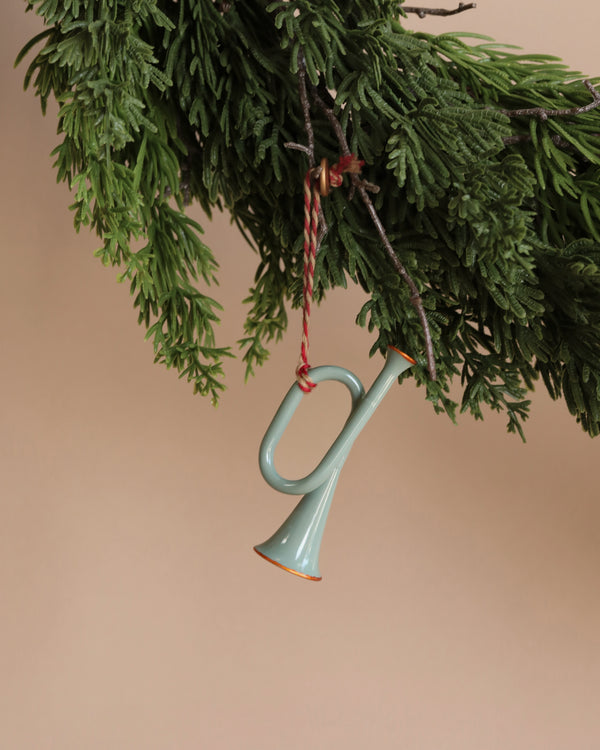 A small Maileg Metal ornament, the Mint Trumpet, featuring blue tones and intricate hand-painted details, dangles from a lush green evergreen branch with a red-and-white string, adding an artistic flair to the festive display against a light brown background.