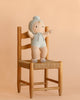 The Cuddle + Kind My First Baby (Bluebell), a handmade baby doll in a light blue outfit, stands on a wooden chair with a woven seat against a beige background.