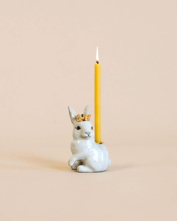 A whimsical hand painted porcelain candle holder shaped like a Royal White Rabbit Cake Topper adorned with a golden crown, holding a lit yellow candle against a soft beige background.