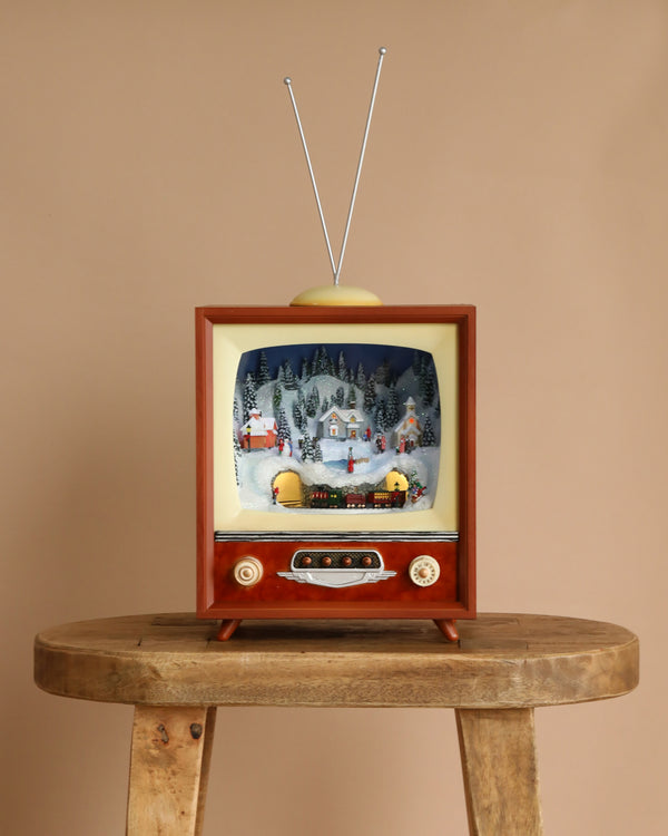 The Christmas Music TV - Town with Rotating Train features a vintage TV with antennas displaying a festive miniature village and moving train inside its screen, set on a wooden stool—perfect for charming holiday decor.