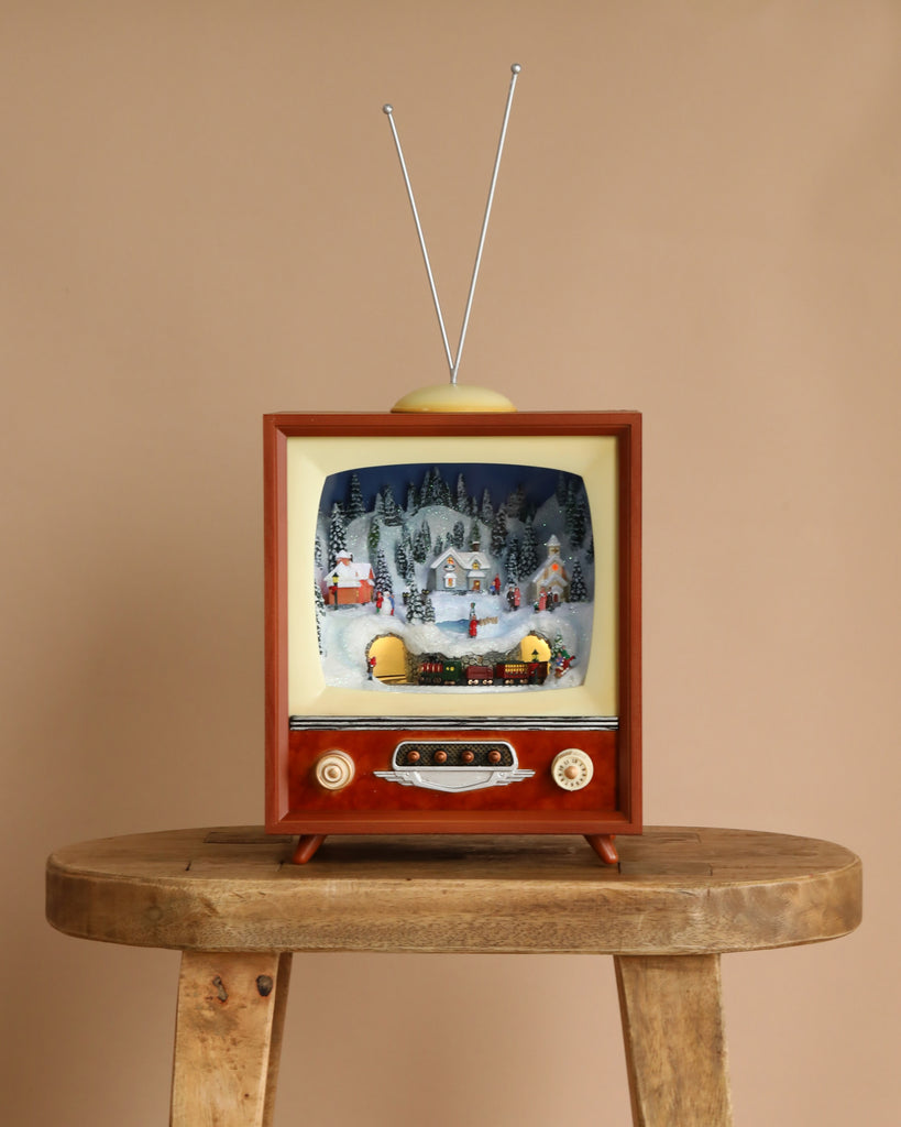 The Christmas Music TV - Town with Rotating Train features a vintage TV with antennas displaying a festive miniature village and moving train inside its screen, set on a wooden stool—perfect for charming holiday decor.
