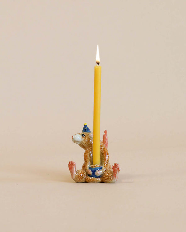 A whimsical Mouse Cake Topper featuring a small, brown squirrel sitting upright, holding a tall, burning yellow candle on its head, against a soft beige background.