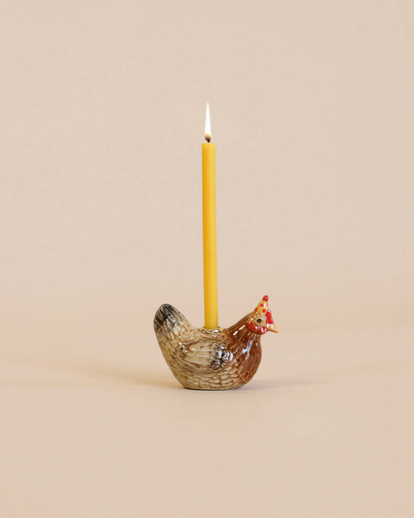 A lit yellow candle stands upright in a Rooster Cake Topper holder, set against a soft beige background. This hand-painted porcelain holder makes it not only functional but also a collectible keepsake.