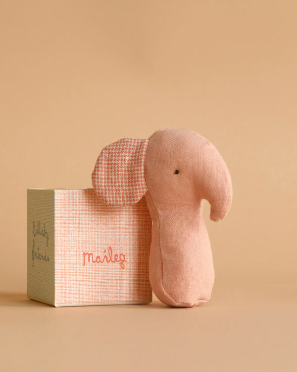 A small, pale pink stuffed elephant crafted from soft fabrics, with one ear featuring unique prints and the other plain, stands next to a box labeled "Maileg Lullaby Friend Rattles, Elephant - Rose." The background is a solid, warm beige color.