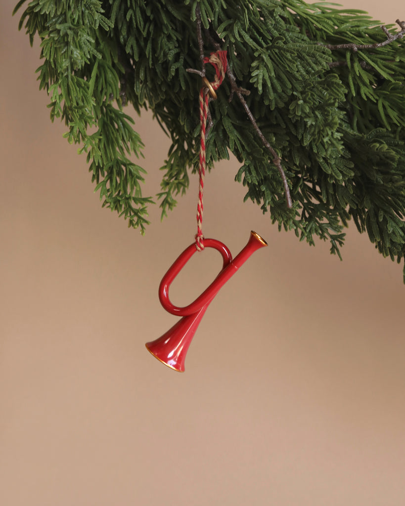 A Maileg Metal ornament, Trumpet - Red, artfully hangs from the branch of a green evergreen tree against a beige background.