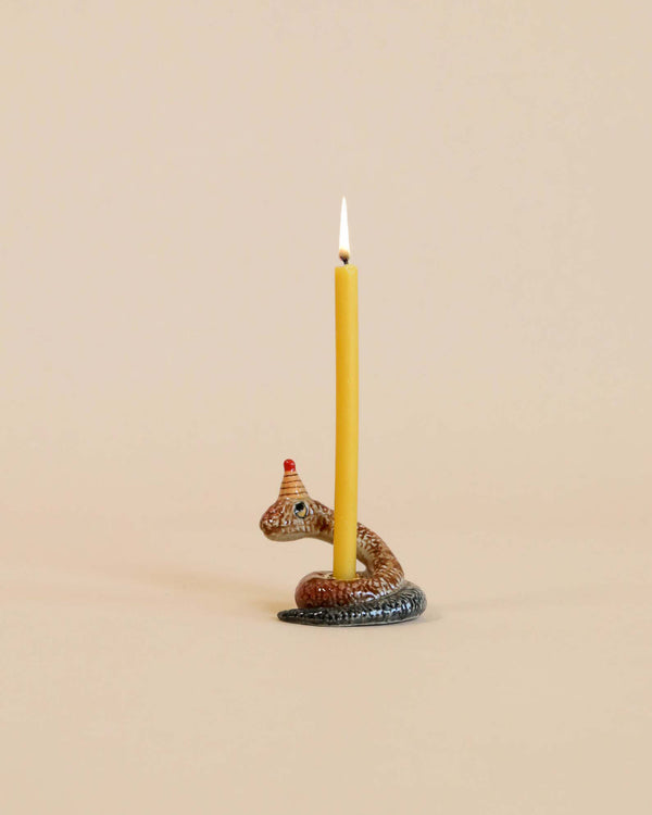 A lit yellow candle stands upright, anchored in a small, decorative holder shaped like a Snake Cake Topper, hand painted and set against a plain beige background.