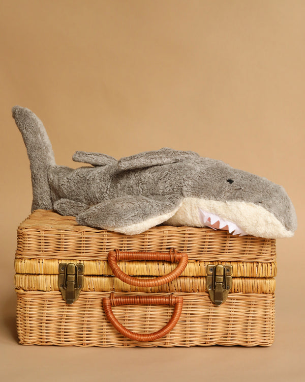 The Senger Naturwelt Cuddly Animal - Shark, a small gray plush with a white underside and soft teeth, rests atop a closed wicker suitcase with leather handles and brass clasps against a beige background.