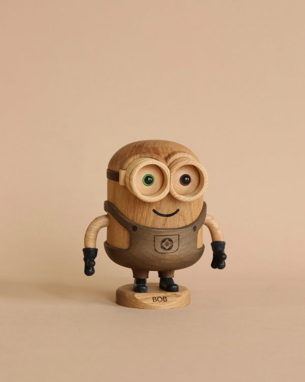 A wooden figurine of Boyhood Minion Bob, with big green eyes, a rounded body, and small arms, standing against a neutral beige background.