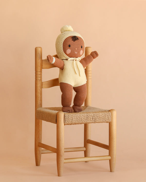 A Cuddle + Kind My First Baby (Buttercup) brown plush hand-knit doll, dressed in a cream bonnet and romper, stands with arms outstretched on a woven wooden chair against a beige background.