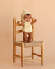 A Cuddle + Kind My First Baby (Buttercup) brown plush hand-knit doll, dressed in a cream bonnet and romper, stands with arms outstretched on a woven wooden chair against a beige background.