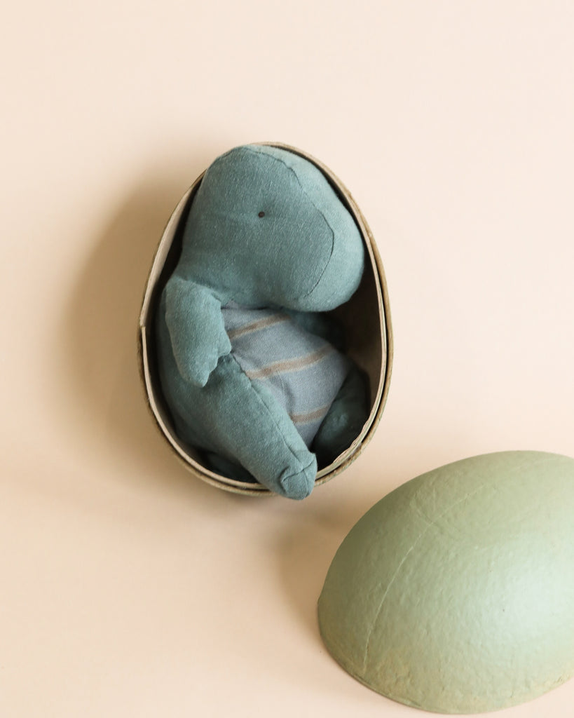 A Maileg | Small Gantosaurus toy curled up inside a round, open paper mache egg against a light beige background, with the top half of the egg lying beside it.