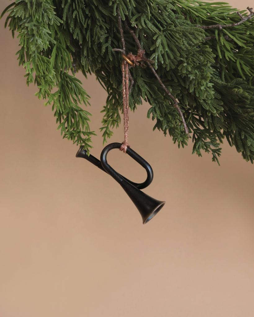 An anthracite trumpet metal ornament from Maileg, featuring handpainted details, hangs from a green conifer branch against a neutral beige background.