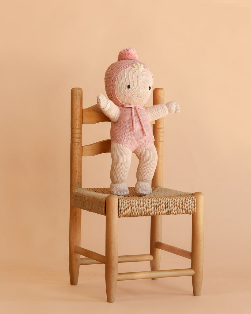 The Cuddle + Kind My First Baby (Rose), a hand-knit baby doll dressed in a pink outfit and hat and crafted from natural cotton yarn, stands on a wooden chair with a woven seat against a plain beige background.