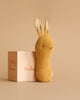 A Maileg Lullaby Friend Rattles, Bunny - Dusty Yellow stands next to a small textured beige gift box. The toy features stitched facial details and upright ears. The box, with red text reading "Lullaby Friends Maileg," enhances the monochromatic backdrop in a matching light brown hue.