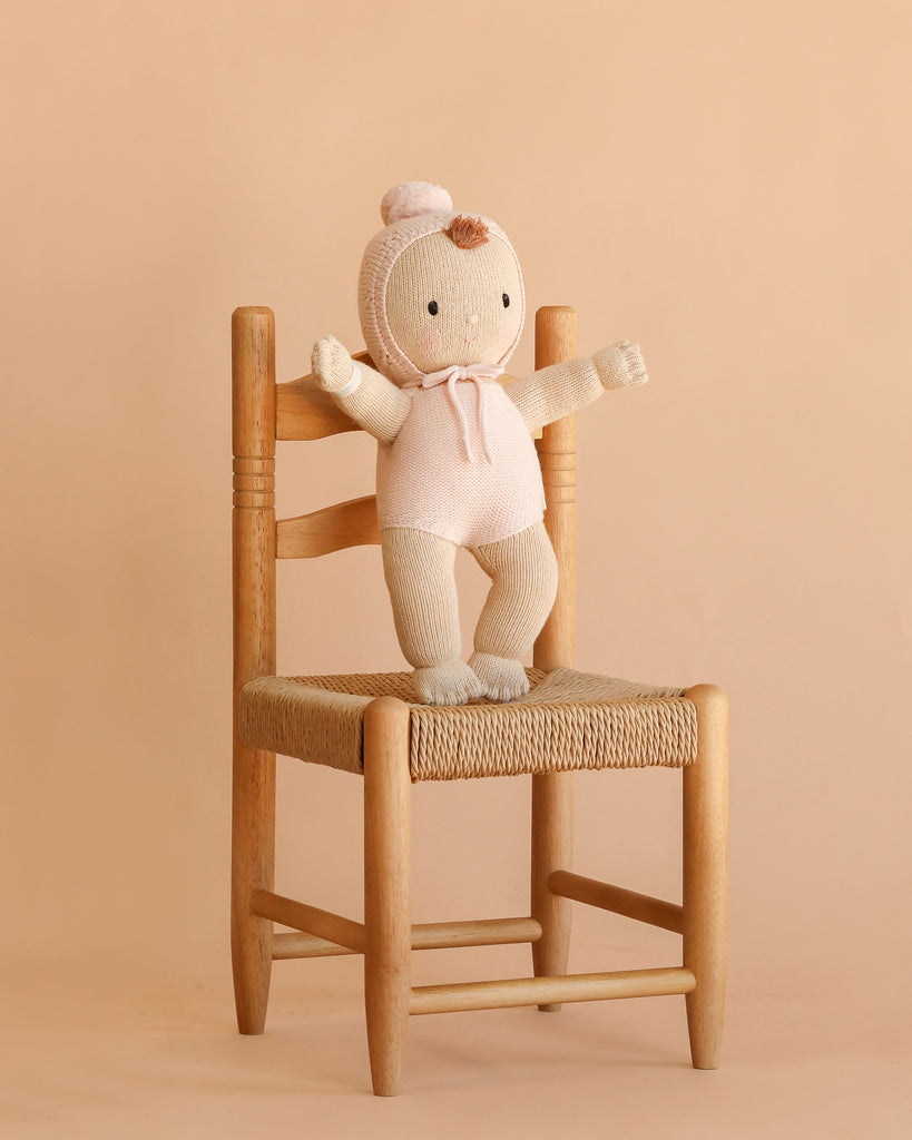 A Cuddle + Kind My First Baby (Blush) doll, hand-knit from natural cotton yarn and dressed in a blush bonnet and outfit, sits on a light wooden chair with a woven seat against a beige background. This fair trade treasure brings warmth and charm to any nursery.