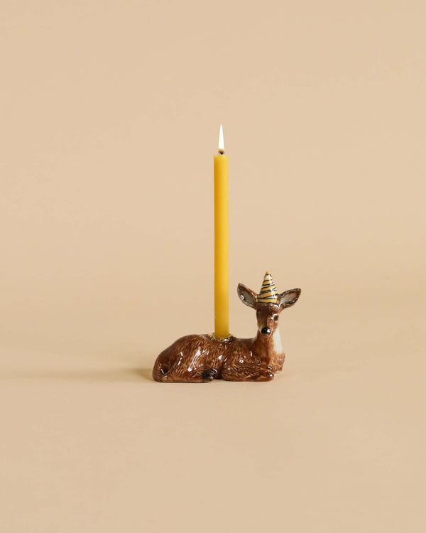 A Deer Cake Topper with a small party hat holds a lit yellow taper candle, set against a plain beige background.