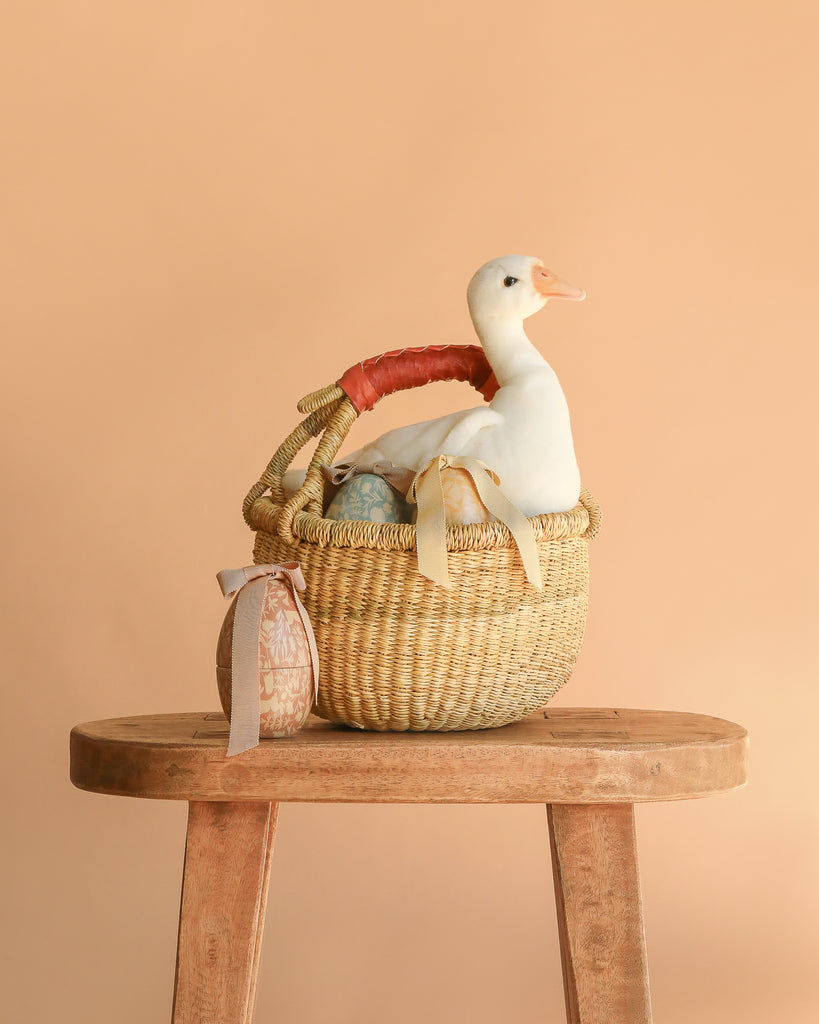 The Easter Basket Gift Set features a young duck stuffed animal in a handmade basket with ribbons, displayed on a wooden stool against a plain beige background.