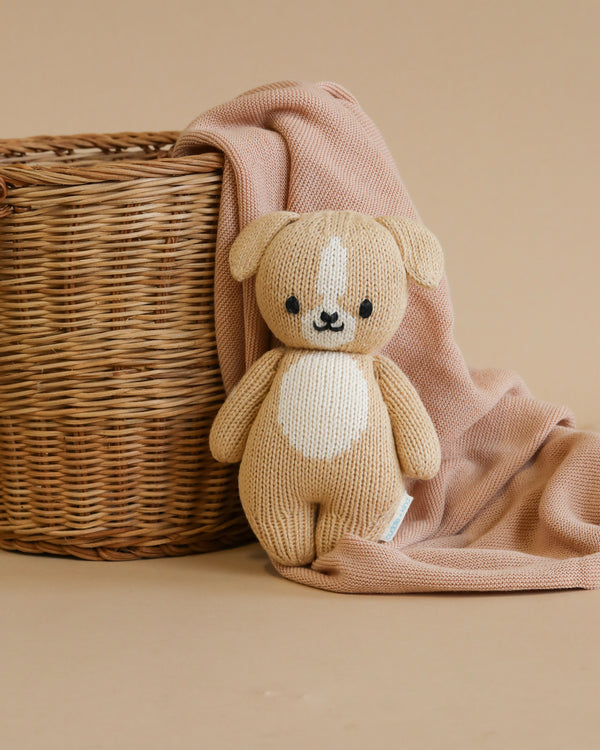 A small, hand-knit Cuddle + Kind Baby Puppy crafted from Peruvian cotton yarn sits in front of a wicker basket, partially draped with a soft pink blanket against a neutral backdrop.