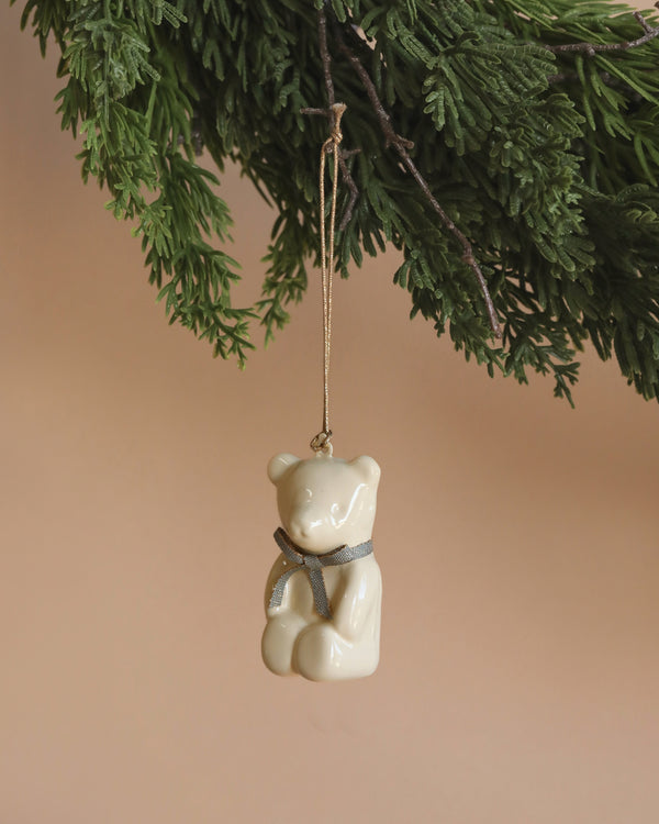 A Maileg metal ornament, featuring an off white and blue teddy bear design, is hanging by a ribbon from a green branch, set against a plain beige background.