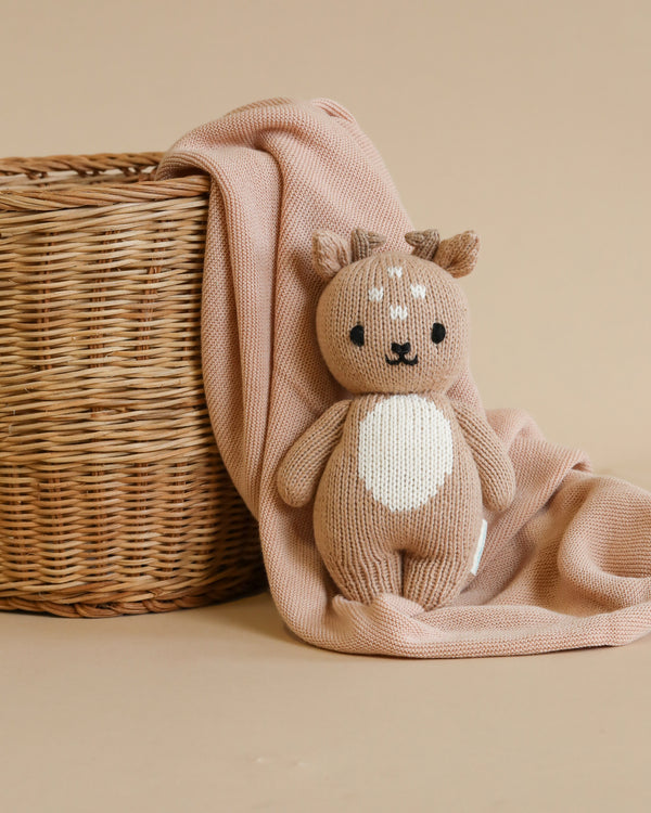 A Cuddle + Kind Baby Fawn hand-knit plush toy deer sits beside a wicker basket, partially draped with a soft pink blanket against a beige background.