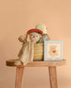 The Easter Basket Gift Set, featuring handmade baby toys and a beige blanket in a woven basket on a wooden stool, is displayed with a floral teddy bear card against a plain peach background.