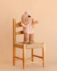 The Cuddle + Kind My First Baby (Peach) doll, dressed in a pink outfit and hat and handmade from natural cotton yarn, is displayed standing on a light wooden chair with a woven seat, set against a beige background.