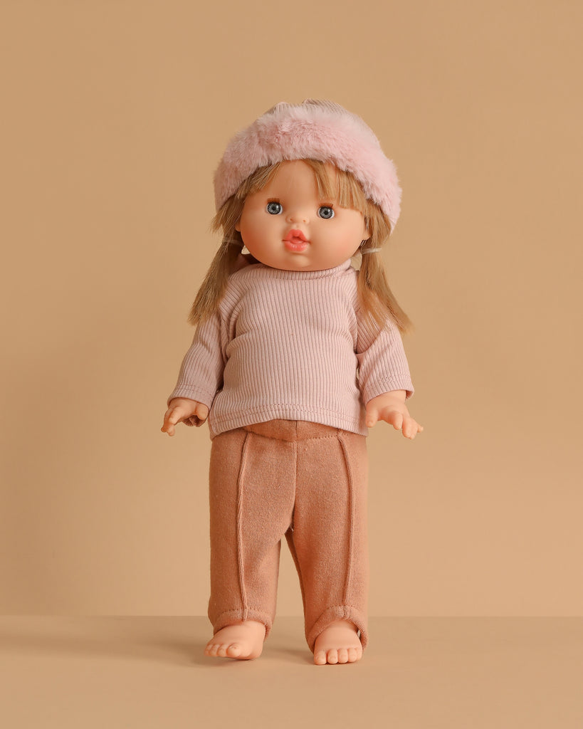 The Minikane Standing Doll (14") - Yzé With Clothing features long blonde hair, blue eyes, and rosy cheeks. Yzé is dressed in a pink long-sleeve top, peach pants, and a fluffy pink hat, standing on a beige surface against a beige background.
