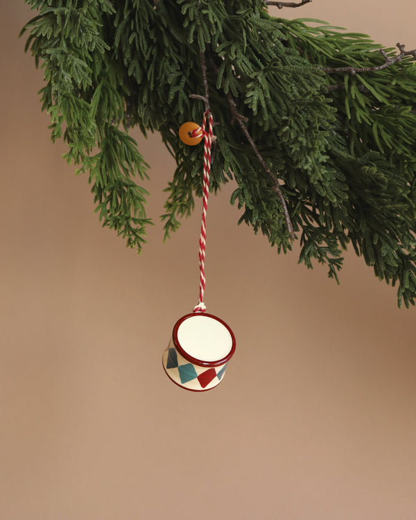 The Maileg Metal Ornament, Small Drum - Classic Red, features hand-painted details in a red and blue harlequin pattern and hangs from a red and white string on a fir branch. The neutral beige background enhances this festive decor, combining nostalgic charm with artisanal elegance.