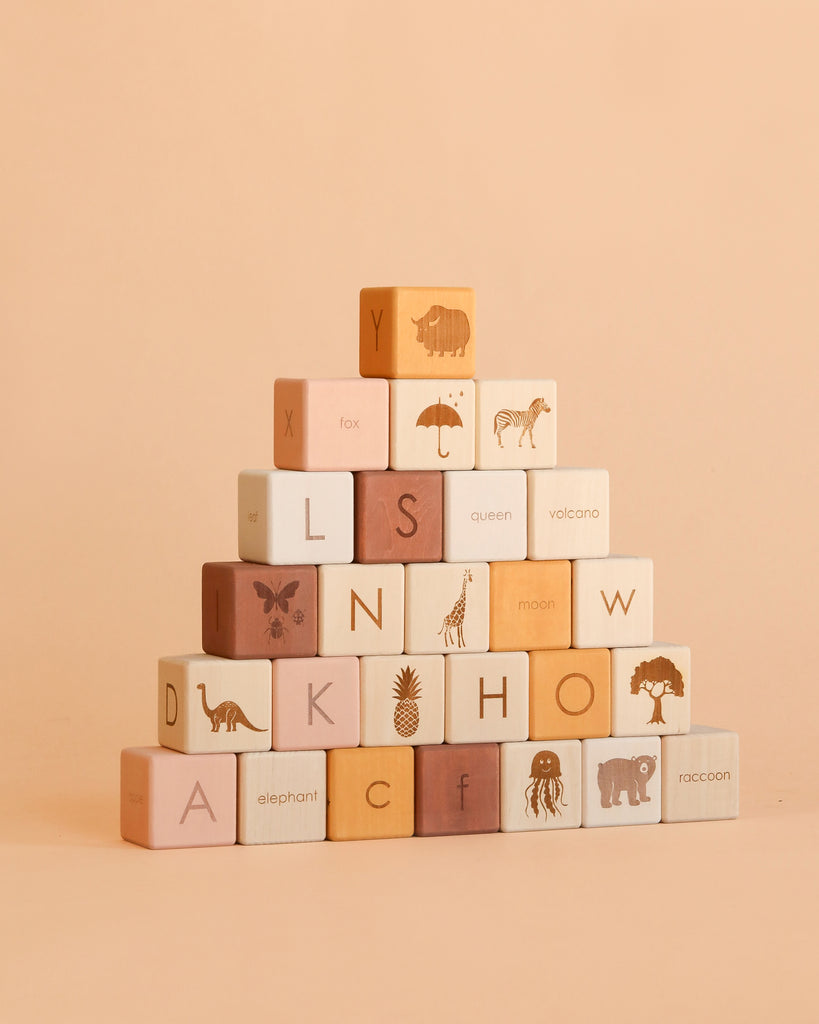 A pyramid of pastel rose-colored Sabo Concept Alphabet Wooden Blocks, meticulously crafted from natural linden wood and finished with non-toxic paint. Each block features letters, animals, and objects illustrated on its surface—displayed against a beige background in the brand’s signature style.