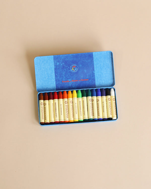 A tin case of sixteen colourful, non-toxic Stockmar wax stick crayons neatly arranged by color spectrum in a blue cardboard box with a decorative lid, placed on a light brown surface.
