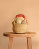 The Easter Basket Gift Set, handcrafted from elephant grass with a red handle, sits on a wooden stool filled with pastel eggs and a Maileg Easter Egg, set against a plain beige background.