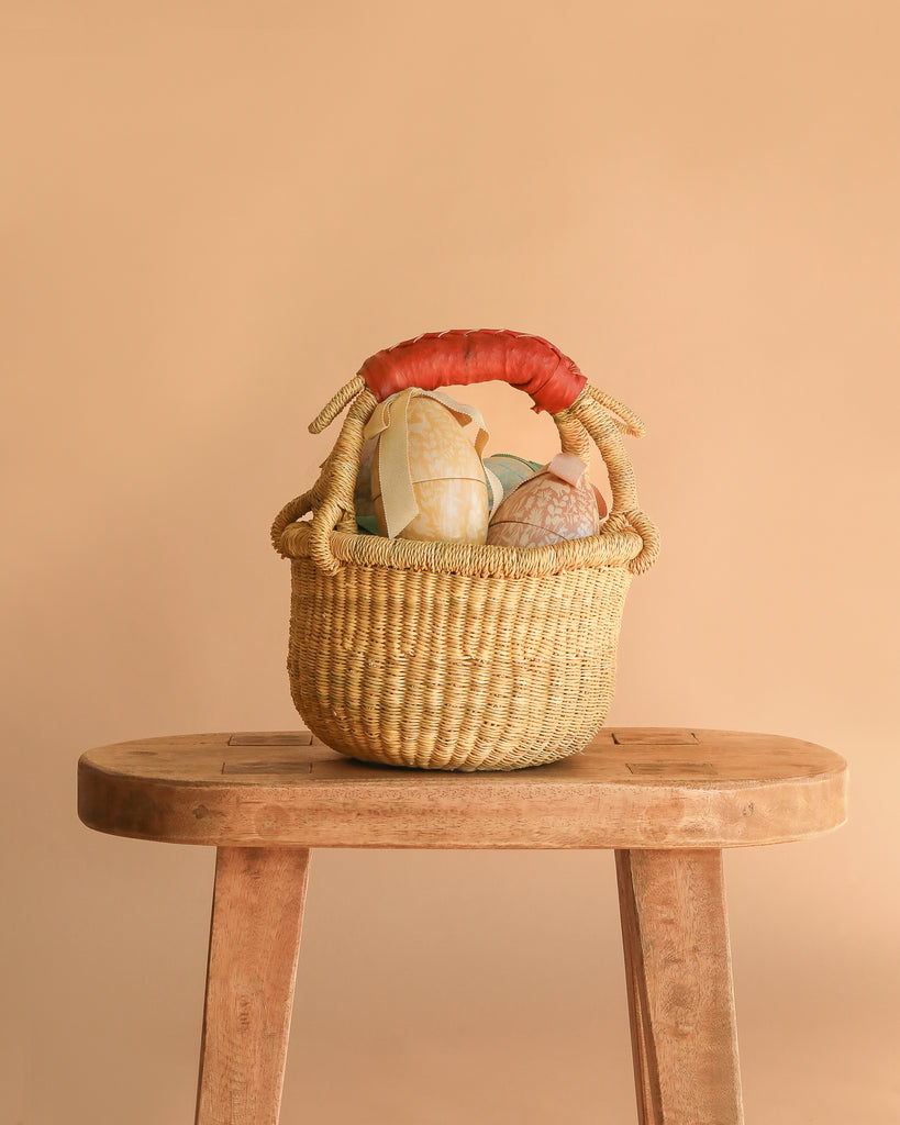 The Easter Basket Gift Set, handcrafted from elephant grass with a red handle, sits on a wooden stool filled with pastel eggs and a Maileg Easter Egg, set against a plain beige background.