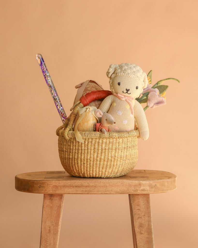 The Easter Basket Gift Set sits on a wooden stool and includes a Cuddle + Kind Lucy the Lamb doll, plush bunny, fairy wand, fabric carrot, Maileg Easter Egg, and artificial flowers against a beige background.