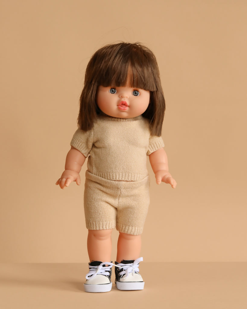 The Minikane Standing Doll (14") - Chléa With Clothing features straight brown hair and comes dressed in a beige knit outfit with white sneakers with black accents, posed against a beige background. Made in Spain and anatomically correct.
