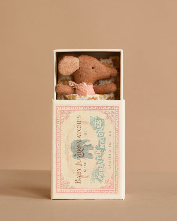 The Maileg Sleepy Wakey Baby Mouse in Matchbox - Rose features a baby mouse in a pink dress resting inside a vintage-style matchbox, creating a charming display against a beige background.