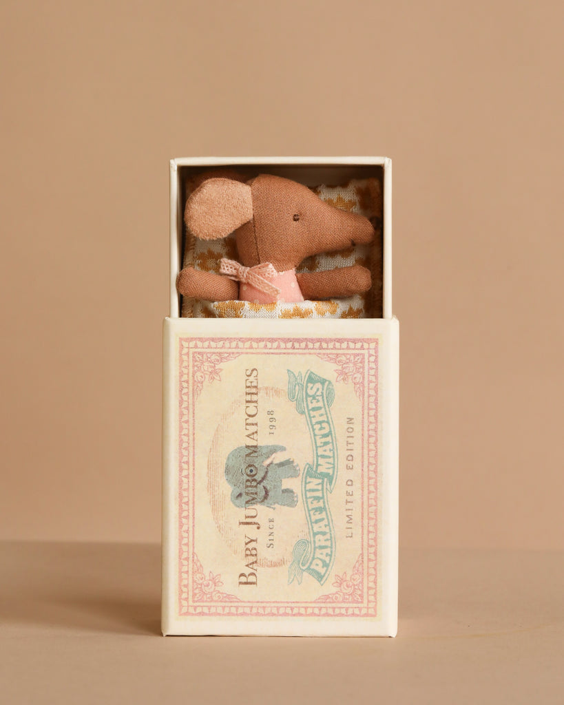 The Maileg Sleepy Wakey Baby Mouse in Matchbox - Rose features a baby mouse in a pink dress resting inside a vintage-style matchbox, creating a charming display against a beige background.