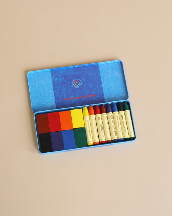 A set of colorful square pastels and Stockmar Wax Crayons Combo Standard Tin Case - 8 Blocks & 8 Sticks Assorted in a metal case arranged neatly next to each other, on a plain beige background. The case lid, visible, has a worn blue cover.