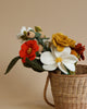 A wicker basket filled with a Felt Flower Bouquet - Autumn, including red, white, and yellow blossoms with green leaves, against a soft beige background.