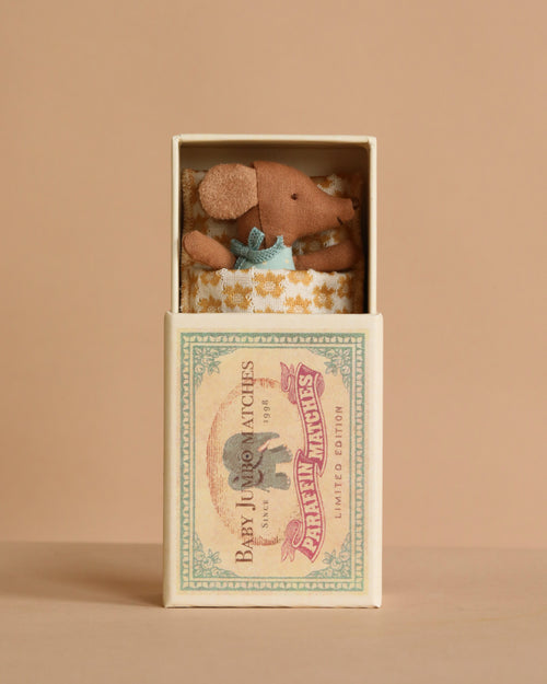 The Maileg Sleepy Wakey Baby Mouse in Matchbox - Blue features a tiny mouse with a blue outfit, nestled in a soft blanket inside a vintage-style matchbox. The background is soft beige.