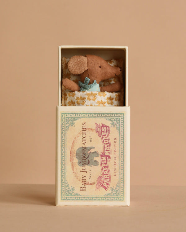 The Maileg Sleepy Wakey Baby Mouse in Matchbox - Blue features a tiny mouse with a blue outfit, nestled in a soft blanket inside a vintage-style matchbox. The background is soft beige.