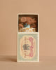 The Maileg Sleepy Wakey Baby Mouse in Matchbox - Blue features a tiny mouse with a blue outfit, nestled in a soft blanket inside a vintage-style matchbox. The background is soft beige.