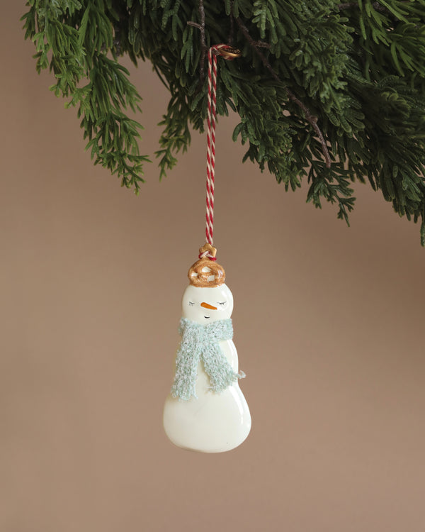 The Maileg Christmas Metal Ornament - 2 Snowman features a charming hand-painted snowman with a light blue scarf and a gold hat, suspended elegantly from a striped red and white string on a green Christmas tree branch against a beige background.