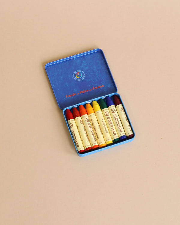 A Stockmar Wax Stick Crayons Waldorf Tin Case containing eight non-toxic colorful crayons arranged in neat rows, displayed against a pale pink background.