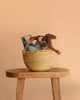 The Easter Basket Gift Set, filled with children's toys like a plush horse, Maileg Cowboy Mouse, fabric doll, and blue ball, rests on a wooden stool against a plain peach-colored background.
