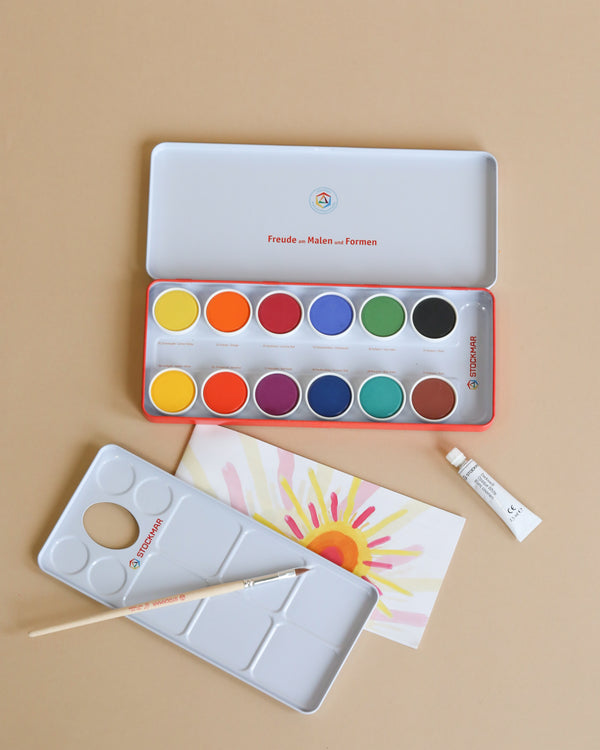 An open Stockmar Opaque Colors - 12 Colors watercolor paint set with various bright colors, two paintbrushes, a small white palette, a tube of white paint, and a paper with colorful paint strokes displayed on.