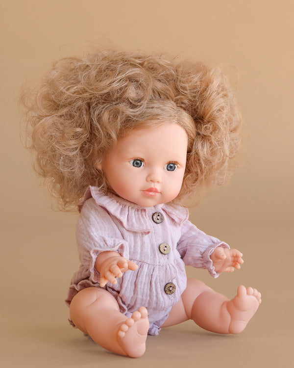 The Minikane Baby Doll (13") - Lola with Clothing features curly hair, blue eyes, and a light mauve ruffled romper with buttons. This anatomically correct doll adds lifelike charm to playtime, pictured against a beige background.