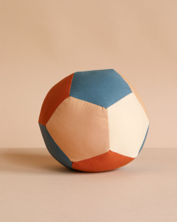 The Fabric Ball - Cobblestone features pentagon-shaped panels in blue, beige, and rust made from soft organic cotton, resting on a neutral surface with a matching beige background.