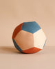 The Fabric Ball - Cobblestone features pentagon-shaped panels in blue, beige, and rust made from soft organic cotton, resting on a neutral surface with a matching beige background.