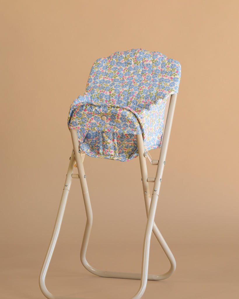 The Konges Sloejd High Chair - Rose Flower is a beige metal foldable doll chair featuring a seat and backrest covered in blue floral-patterned fabric, shown against a plain beige background.